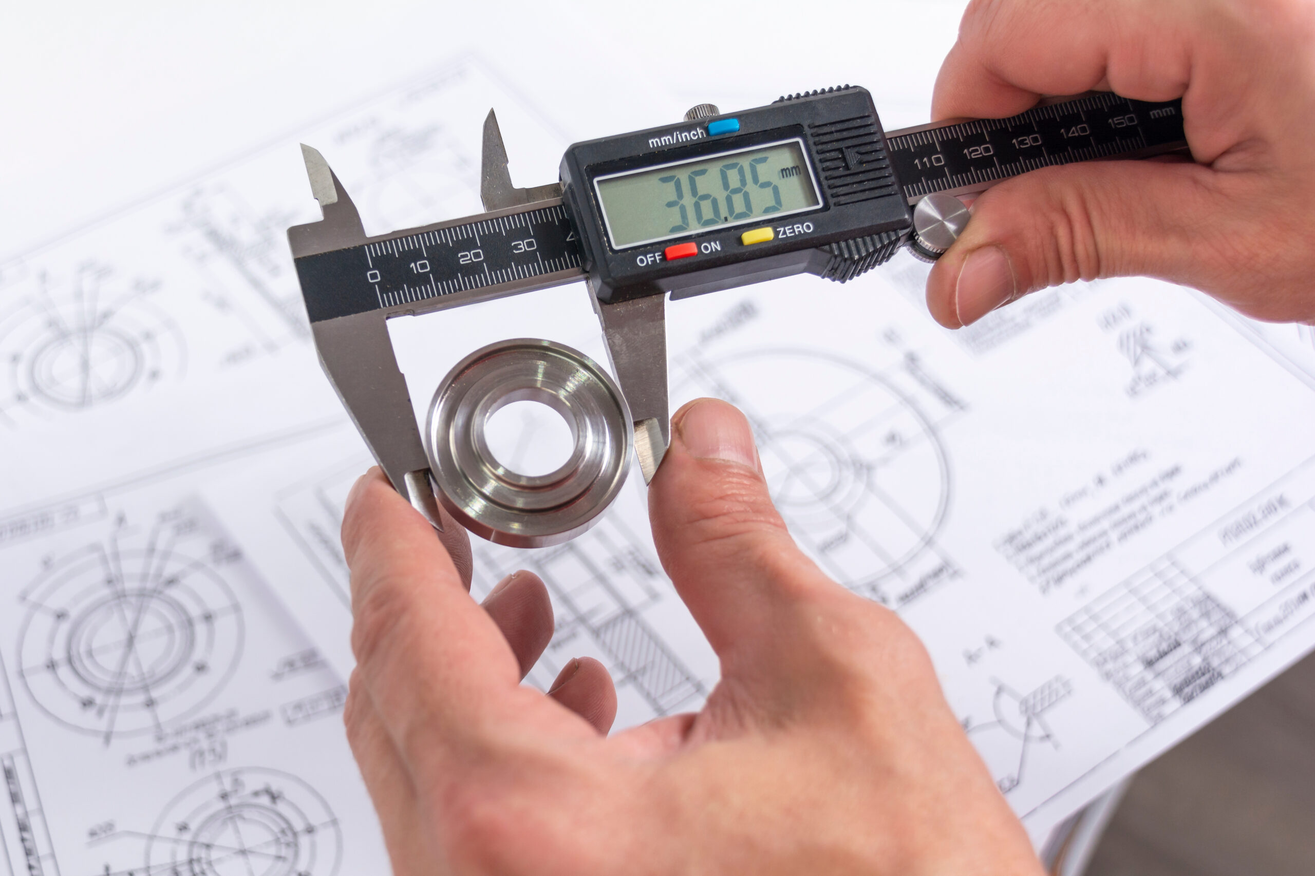 Hands of an engineer measures a metal part with a digital vernier caliper against the background of technical drawings. Quality control of parts machined on a lathe.